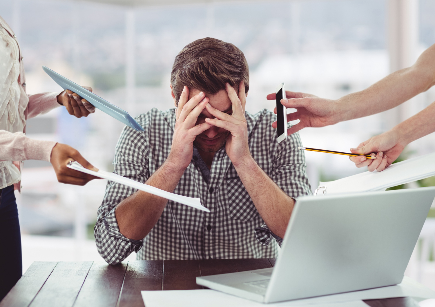 Homem sentado à mesa de trabalho com as mãos na cabeça, cercado por colegas oferecendo documentos e telefone, representando a necessidade de gerenciamento do estresse no ambiente profissional.