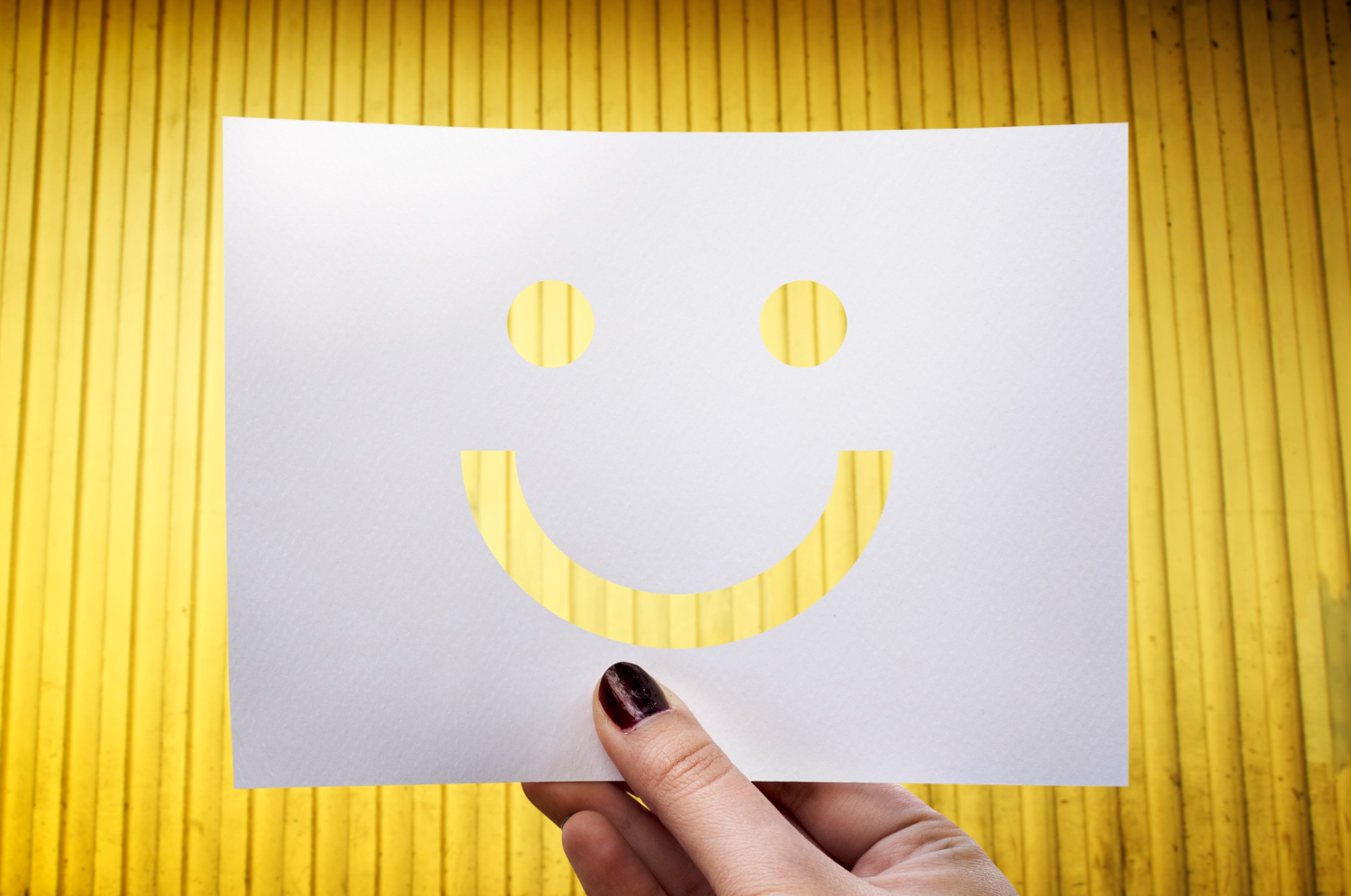 Mão segurando um papel branco recortado em formato de rosto sorridente, com fundo amarelo vibrante, simbolizando positividade e otimismo.
