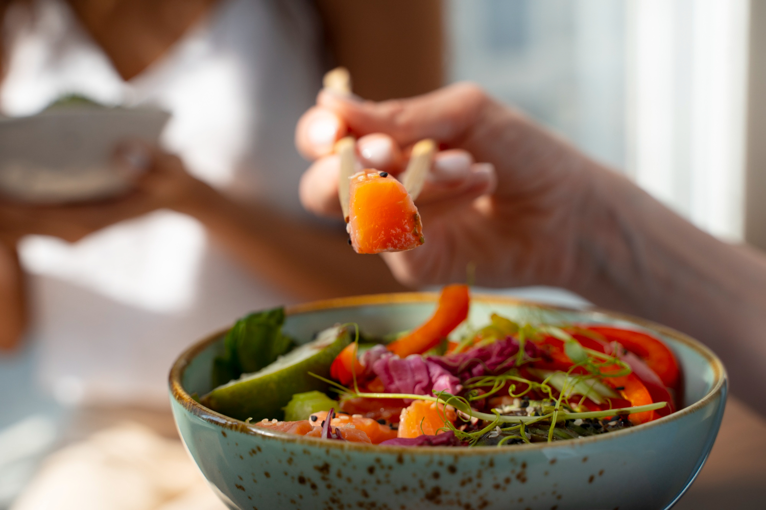 Pessoa segurando palitinhos com um pedaço de salmão fresco sobre uma tigela colorida de legumes e verduras, representando uma refeição saudável e equilibrada.