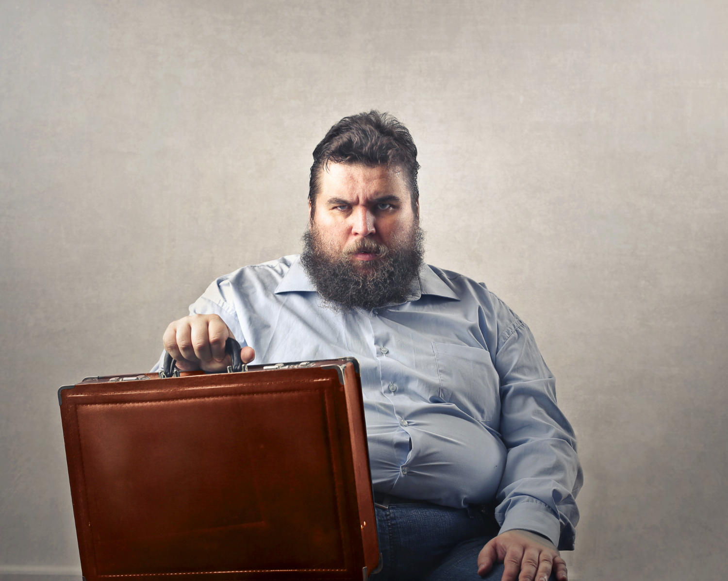Homem de barba e aparência séria, vestindo camisa social azul clara, sentado com uma pasta de couro nas mãos, representando o tema profissões com o maior ganho de peso.