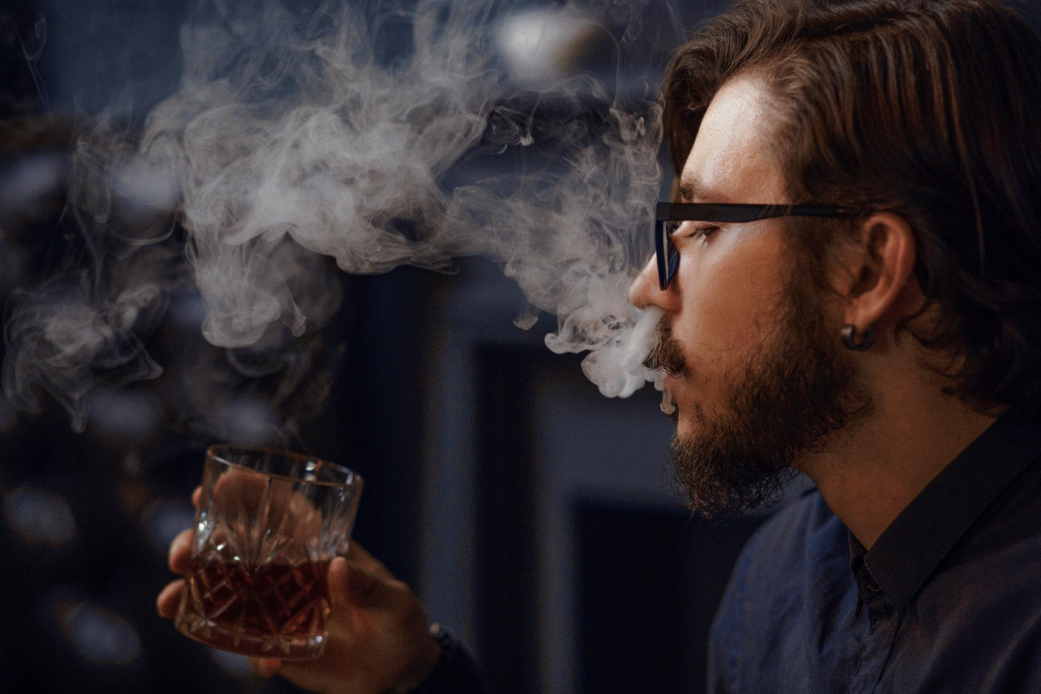 Homem soltando fumaça enquanto segura um copo com bebida alcoólica, representando hábitos prejudiciais à saúde que devem ser evitados.