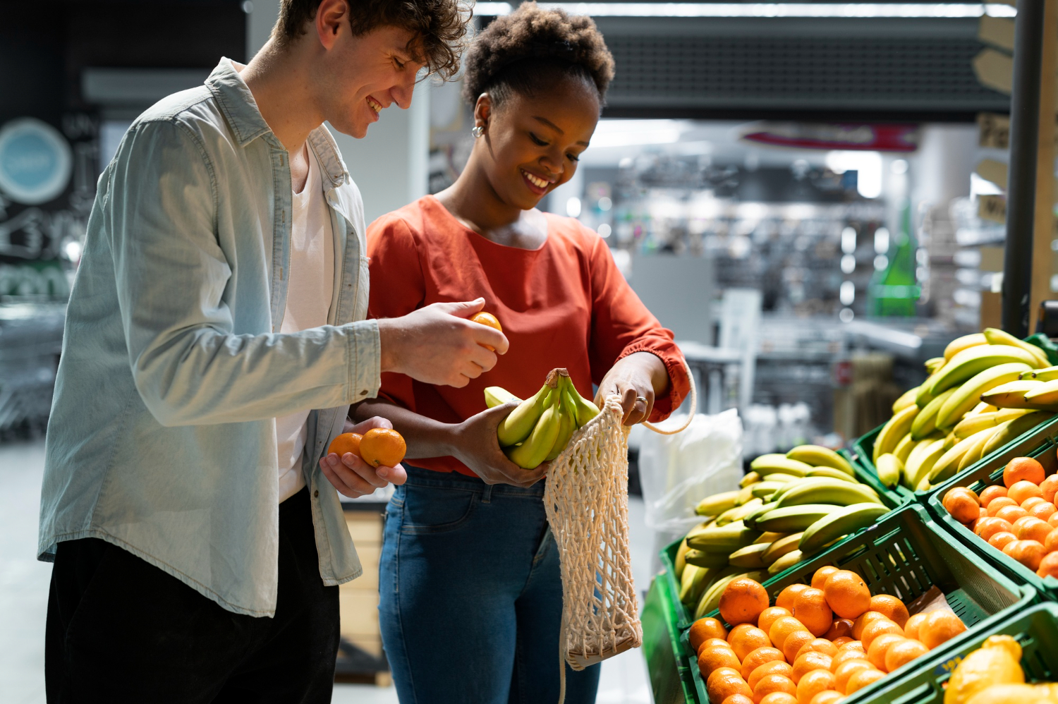 Casal sorridente escolhendo bananas e tangerinas frescas na seção de frutas de um supermercado, colocando os produtos em uma sacola reutilizável.
