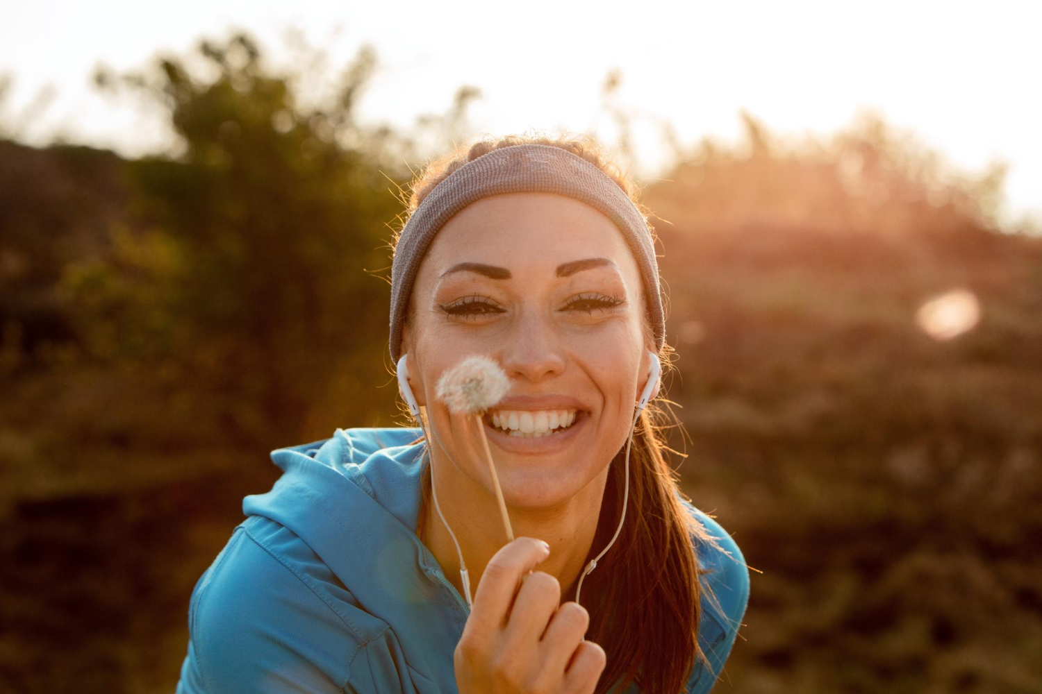 Mulher sorridente ao ar livre, usando roupas esportivas e segurando uma flor de dente-de-leão, aproveitando o momento de bem-estar sob a luz do sol.