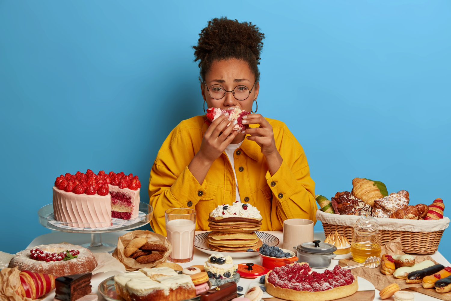 Mulher sentada à mesa repleta de doces e bolos, com expressão triste enquanto come, simbolizando o impacto emocional da ansiedade alimentar.