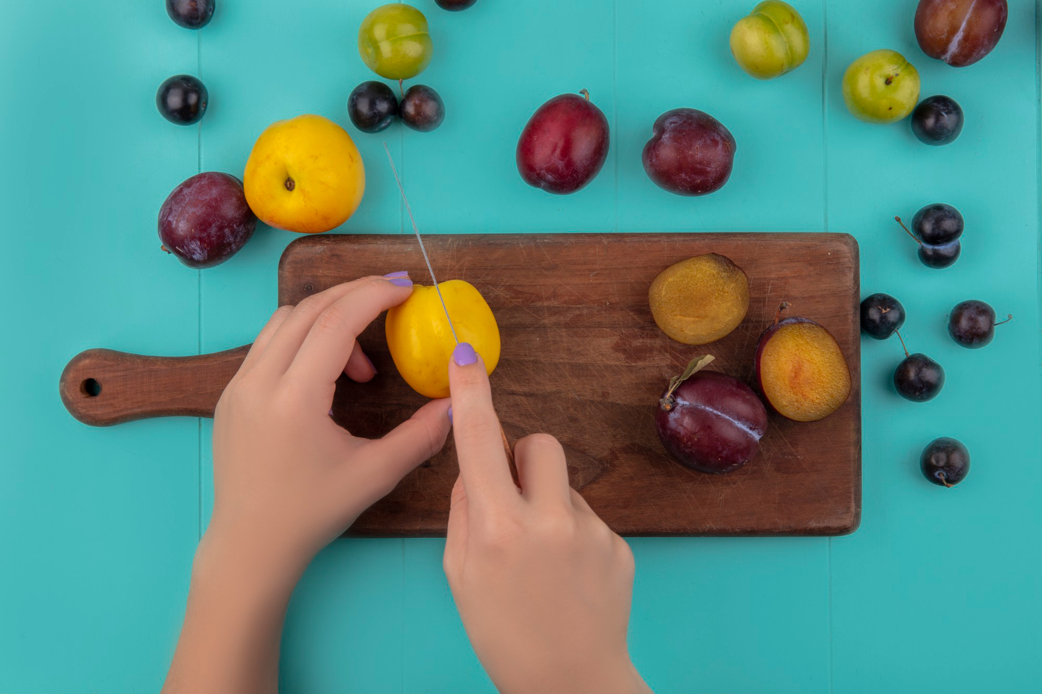 Mãos cortando uma fruta amarela sobre uma tábua de madeira, cercada por diversas ameixas e frutas coloridas, representando a diferença entre beliscar e fracionar as refeições.
